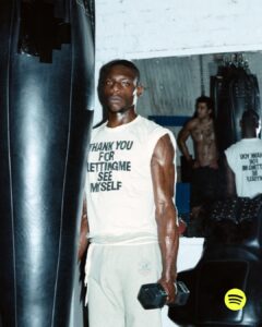 A man with a serious expression stands in a gym, holding a dumbbell. He wears a sleeveless shirt and sweatpants. Sweat glistens on his muscular arms, indicating a workout. Behind him, a punching bag and a mirror reflect another person. The setting suggests a focused, intense atmosphere typical of a space dedicated to physical training.