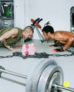 Two people face each other doing push-ups on a gym floor, with a plush pink toy resting on a chain between them. One person has a tattooed arm and wears a sleeveless top, while the other is shirtless, wearing fingerless gloves. A barbell lies in the foreground, and gym equipment is visible in the background.