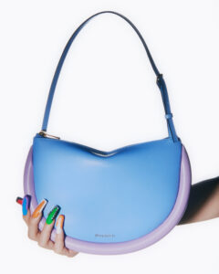 A crescent-shaped blue handbag with a purple border features a single strap. A hand with long, colorful nails in shades of blue, orange, green, and white holds the bag. The design is modern and vibrant, showcasing a playful contrast. The nails include unique patterns and small embellishments, enhancing the overall artistic and whimsical appeal.