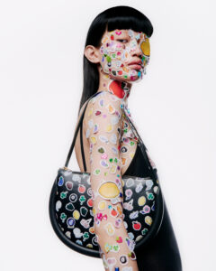 A person stands confidently, adorned with a profusion of colorful stickers across their face and arm. Sporting sleek, straight hair, they wear a stylish black ensemble. A black bag with a similar sticker motif hangs from their shoulder, complementing the vibrant expression of individuality and creativity, against a stark white backdrop that highlights the design.