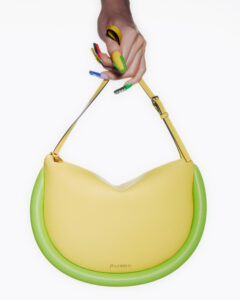 A hand with vibrantly colored nails holds a stylish, crescent-shaped bag. The bag features a yellow body accented with a green, rounded base and an adjustable strap. The nails are painted in bold hues of blue, red, green, yellow, and black, adding a pop of color and flair to the scene, enhancing its modern aesthetic.
