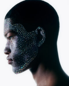 A person is depicted in profile with a bald head against a white background. Their face is intricately adorned with sparkling, multicolored gemstones, creating a striking and artistic appearance. The stones cover the forehead, around the eyes, cheeks, and chin, reflecting light and adding a sense of depth and texture to the skin's surface.