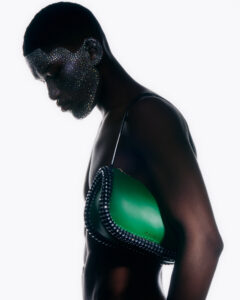 A figure with dark skin gazes downward, their face adorned with shimmering beads. They wear an elegant green accessory with a black strap that is accentuated by intricate detailing along the edges. The minimalist aesthetic emphasizes the contours of the body, offering a bold contrast between the glossy accessory and the textured embellishments on the figure's face.