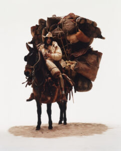 A person in a cowboy hat sits astride a heavily laden horse, surrounded by sand. The horse is burdened with numerous bags and rolled-up items, appearing ready for a long journey. The rider is dressed warmly in a puffer jacket and boots, looking directly ahead. The scene conveys a sense of rugged determination and adventure.