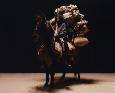 A solitary figure rides a donkey, enveloped in a dimly lit scene. The rider wears layered clothing and holds the reins with a determined expression. The donkey is heavily laden with assorted bags, blankets, and parcels, evoking a sense of a long journey. The ground beneath is bare, contrasting against the dark backdrop, enhancing the overall atmosphere.