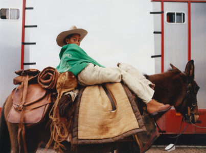 A child in a large white cowboy hat and green jacket sits atop a mule. The mule is equipped with a brown leather saddle and rolled blanket. The child gazes back with a relaxed expression. Their pants are beige, and boots are brown. In the background, a red and gray vehicle with small windows is visible.