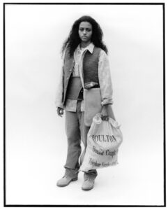 A person with curly hair stands against a plain background, wearing layers of casual clothing including a textured vest, a plaid shirt, and rolled-up trousers. They carry a large sack-like bag with a strap over their shoulder. Their expression is neutral, and footwear consists of casual, laced shoes. The setting is minimalist and monochrome.
