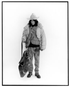 A person stands against a plain background, wearing a cowboy hat, puffy jacket, and layered clothing underneath. They hold a large, worn bag in their right hand. Their attire includes loose-fitting pants and casual shoes. The monochrome setting highlights the rugged, utilitarian style, evoking a sense of adventure or journey in a minimalist, yet striking, manner.