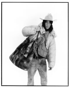 A person stands confidently, wearing a cowboy hat and a puffy jacket. Their expression is calm, with long hair cascading past their shoulders. Slung casually over their shoulder is a large, textured bag. The scene is minimalistic, focused on the individual’s unique style, featuring layered clothing and a relaxed, yet intriguing pose on a plain background.