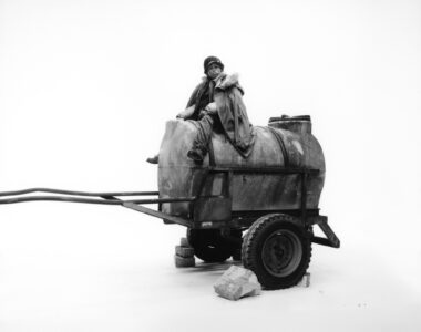 A person wearing a heavy coat and hat sits atop a cylindrical metal tank on a wheeled cart. The cart is supported by bricks and has large, sturdy wheels. The overall scene is minimalistic with a stark background, emphasizing the person's contemplative expression. The industrial, weathered look of the cart contrasts with the simple, clean setting.