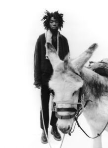 A black-and-white composition features a person with distinct, textured hair, wearing a layered outfit and scarf. They stand calmly holding a rope tethered to a donkey. The donkey, equipped with a harness and burdened with a pack, faces the viewer slightly to the side. The background is minimalist, emphasizing the subjects and their expressions.