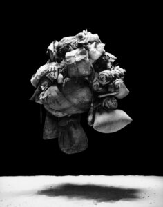 A complex, floating assemblage of various items like bags, cloth, and rolled fabrics displays a rich texture against a stark, black background. Shadows drift beneath the mass, creating a sense of depth and weightlessness. The monochromatic palette emphasizes the intricate details and contours of the objects, evoking a feeling of organized chaos and suspended animation.