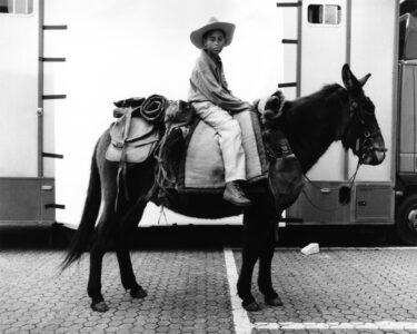 A young person wearing a cowboy hat sits confidently atop a saddled mule. Their attire includes a long-sleeved shirt and boots. The scene is monochromatic, with a backdrop of trailers and a paved surface, evoking a rustic or theatrical setting. The mule is equipped with various bags, suggesting a journey or role in a staged production.