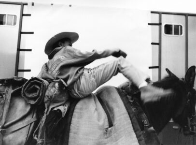 A person wearing a cowboy hat is mounting a horse with their body angled sideways. The horse, adorned with a padded saddle, appears calm during the action. The backdrop consists of a structure with rectangular shapes, adding an industrial touch to the scene. The image captures a dynamic moment, emphasizing movement and the distinctive attire associated with the activity.