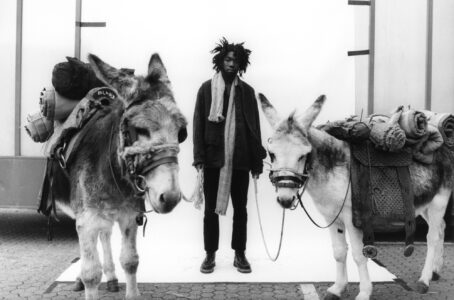 A person with distinctive hair stands between two donkeys, holding their reins. The donkeys are equipped with saddles carrying rolled mats, suggesting they are ready for a journey. The scene is set against a plain backdrop, highlighting the contrast between the individuals and their environment. The monochrome tones give a timeless, classic feel to the composition.
