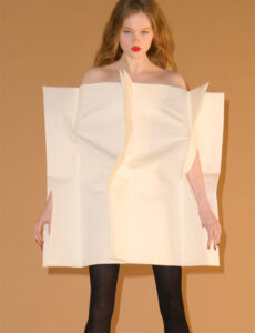 A person stands against a warm beige background, wearing a unique, structured off-white outfit resembling folded paper. Their hair is long and wavy, styled naturally. They have striking red lipstick, adding a pop of color to the composition. Black stockings provide contrast to the outfit, creating a bold, avant-garde fashion statement with a minimalist and artistic appeal.
