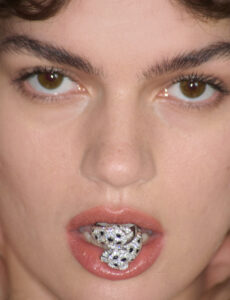 A close-up shows a person with short, dark hair and expressive brown eyes gazing directly ahead. Their lips are slightly parted, revealing a sparkling, jewel-encrusted panther motif nestled inside their mouth. The skin appears smooth and flawless, with a focus on the jewelry's intricate details, enhancing the overall captivating and bold composition.