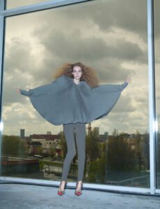A person with voluminous, curly hair stands against a large window, wearing a flowing, pleated gray top and fitted gray leggings. The dramatic top extends with outstretched arms, creating a wing-like effect. Below, red high heels contrast with the muted outfit. Reflections show an urban landscape outside, under a sky filled with clouds.