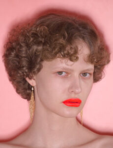 A person with curly brown hair gazes directly with a neutral expression. Their lips are strikingly colored in vivid orange, contrasting with the softer hues around them. Gold dangling earrings frame the face, adding elegance. The backdrop consists of a soft pink tone, enhancing the warm ambiance and highlighting their facial features and accessories harmoniously.