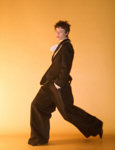 A person with curly hair is striking a dynamic pose, wearing an oversized black suit against a warm, orange backdrop. The suit features a wide-legged style and a large, white accessory near the collar. One hand is tucked into a pocket, enhancing the relaxed yet fashionable demeanor. The lighting creates a soft, golden hue, highlighting the outfit's texture.