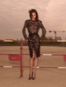 A person stands in front of a blurred industrial background with overcast skies. They are dressed in an intricately patterned black lace outfit featuring a long-sleeved top and a fitted skirt with a knee-high slit. Their hair is tousled by the wind, adding to the dynamic feel. Red and white railings add contrast to the scene.