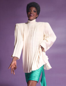 A person joyfully poses against a soft purple backdrop. They wear a flowing, pleated cream blouse with exaggerated shoulders, paired with a vibrant green, ruched skirt. Their confident expression, combined with graceful hand gestures, enhances the overall elegance and liveliness. The ensemble captures a blend of modern style and dynamic motion, exuding confidence and charm.