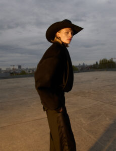 A person stands outdoors, wearing a wide-brimmed hat and dark clothing. Their hands are in the pockets of their trousers. The setting appears urban, with a hint of buildings in the background against a cloudy sky. The scene is bathed in soft, natural light, creating a contrast between the person's attire and the expansive concrete surface.