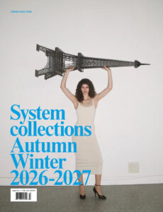 A woman in a fitted cream dress and black heels stands against a sparse interior backdrop, balancing a large architectural structure above her head with poised confidence. Her direct gaze and upright posture create a sense of strength and surreal elegance. The minimalist setting emphasizes scale, contrast, and visual tension, blending fashion, sculpture, and performance into a striking contemporary tableau.