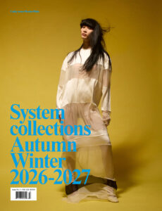 A woman with long, dark hair stands against a mustard-yellow background. She wears a sheer, layered dress with flowing fabric that creates movement. Her pose is relaxed yet confident, with a slight tilt of her head. The lighting casts soft shadows, accentuating the texture and fluidity of the outfit, creating a harmonious and artistic composition.