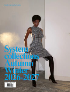 A person with a confident posture sits elegantly on a white chair against a minimalist backdrop. They wear a black and white checkered dress with a high neck and cut-out details, paired with dark tights and shiny black heels. The setting has a soft lighting effect that casts a subtle shadow on a plain wall and concrete floor.