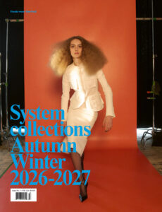 A model with voluminous curly hair strikes a poised, dynamic pose against a vibrant orange backdrop. She wears a fitted white suit, comprising a structured jacket and pencil skirt. Her expression is confident and forward-looking. Studio lights and equipment are visible, adding a behind-the-scenes feel to the setting. The overall tone is sophisticated and fashion-forward.