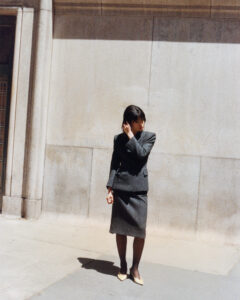 A woman dressed in a dark gray business suit stands on a sunlit sidewalk beside a large stone wall. Her hair is styled neatly, and she appears to be adjusting it with one hand. Wearing dark stockings and light-colored shoes, she gazes away from the camera, casting a subtle shadow on the ground.