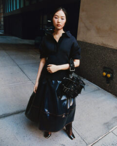 A young woman stands on a city sidewalk, wearing a stylish black outfit featuring a textured skirt and matching top. She carries a unique black handbag with fringe details. Her expression is calm and poised, and her hair is pulled back neatly. The surrounding environment includes concrete pavements with shadows from nearby buildings.