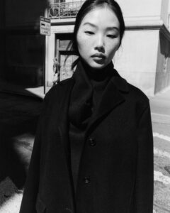 A striking black-and-white composition features a person with long hair and freckles. They wear a dark coat in an urban setting, emphasized by strong shadows and sunlight. The background includes a building and a distant pedestrian, creating a contrast between light and dark. The individual gazes downward, lending an introspective mood to the scene.