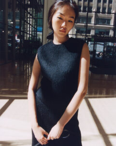 A woman stands indoors, bathed in soft, natural light filtering through large windows. She wears a sleeveless, textured black dress, exuding elegance and confidence. Her expression is calm and poised, with her hands resting gently. Behind her, the modern urban setting features tall glass walls and reflections, adding depth and a contemporary feel to the scene.