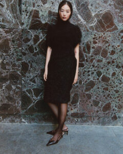 A woman stands against a dark, textured marble wall. She wears a high-neck, short-sleeve black top and a matching knee-length skirt. Her outfit features different textures, with a feather-like appearance on the top. She also sports sheer black stockings and pointed heels with metallic details. Her expression is serene, and her hair is straight and sleek.
