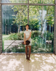 A woman in a white dress sits on a wooden floor, holding a brown bag. She is positioned in front of large glass panels that reveal a lush, sunlit garden with trees and greenery. Soft natural light filters through, casting shadows on the floor. The setting is peaceful and blends the interior with the natural exterior.