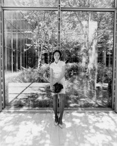 A woman sits on a bench, holding a bag, in a bright and airy space. She wears a light-colored dress and heels. Behind her, large glass panels reveal a lush garden with trees and foliage. Dappled sunlight casts intricate shadows on the floor, creating a serene and contemplative atmosphere. The setting feels modern and peaceful.