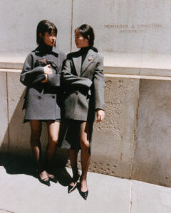 Two women are standing closely on a sunlit street, both dressed in stylish grey coats and black pointed shoes. They are looking at each other with friendly expressions. The background consists of a textured stone wall, casting shadows that add depth. Natural light highlights their features, creating a warm and engaging atmosphere.