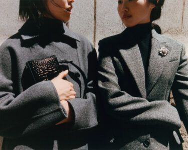 Two individuals are wearing stylish, tailored gray coats. One person holds a small, textured black clutch, while the other wears a dark turtleneck underneath their coat. A decorative brooch embellishes the lapel of the coat on the right. They stand against a light, textured wall, casting subtle shadows. The overall tone is sophisticated and contemporary.