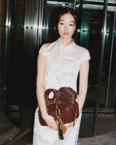 A young woman stands in a sleek, modern setting with a reflective background. She wears a short-sleeved, elegant white dress and holds several brown leather handbags with various textures. Her expression is poised and calm, while the lighting highlights her features softly. The scene exudes a sense of fashion-forward, urban sophistication.