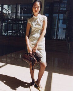 A woman stands in a sunlit interior, casting a shadow on the polished floor. She wears a light, structured dress and holds two handbags. Her shoes are pointed and complement her outfit. The background is urban, visible through large glass windows, suggesting a modern setting. The overall ambiance is warm with contrasting light and shadows.