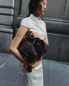 A person wearing a short-sleeved, collared white dress stands in an urban setting. They hold a stylish, dark brown handbag with gold zippers and multiple compartments. Their hair is casually styled, and they seem deep in thought. The surrounding environment is composed of textured stone walls and a concrete pavement, suggesting a city atmosphere.