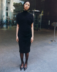 A person stands on a city sidewalk in front of a modern glass building. They're wearing a black outfit consisting of a textured top and knee-length skirt, paired with sheer stockings and shiny pointed shoes. The urban backdrop features reflections and a revolving door, contrasting with their composed and serene expression.