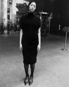 A woman stands confidently on a city sidewalk, wearing a sleek, textured, dark ensemble, accentuated by pointed shoes. The setting features reflective glass buildings and a revolving door in the background. The grayscale tones create a dramatic contrast, highlighting the textures of her attire against the urban architecture, capturing an essence of modern urban sophistication.