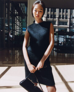 A person stands in a sunlit indoor setting, casting shadows on a polished floor. They wear a sleeveless black dress and hold a black handbag. Large windows in the background reveal an urban exterior with tall buildings. The lighting creates a contrast between the bright sunshine and shaded areas, enhancing the scene’s modern and sophisticated atmosphere.