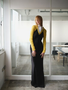 A person stands in a minimalist, modern space with a polished concrete floor and glass wall partition. They wear a sleek black dress under a bright yellow long-sleeve top, with a white collar peeking through. Sunlight filters in from large windows, casting soft shadows. The setting appears to be a blend of industrial and contemporary design.