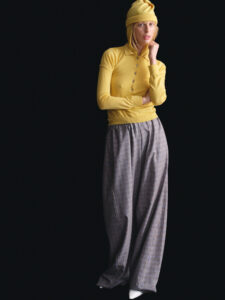 A person stands poised, wearing a bright yellow long-sleeve top and matching hat. Their ensemble is completed with loose-fitting plaid trousers in muted tones. The backdrop is solid black, drawing focus to their contemplative expression and relaxed pose. The contrast of vivid yellow against the dark background creates a striking visual effect.