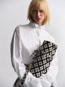 A person with blonde hair is standing against a light background, wearing an oversized white shirt. They are holding a rolled-up, plaid-patterned fabric in black and white. The person's expression is neutral, and the lighting softly illuminates their features. The overall mood is minimalist, with a focus on texture and style.