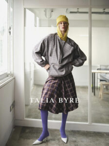 A person stands confidently in a modern interior. They wear a yellow knit hat and a gray, oversized plaid jacket paired with a brown plaid skirt. Vibrant purple tights contrast with white pointed heels. The lighting is soft and natural, entering through large windows, illuminating the minimalist room adorned with clean lines and neutral tones.